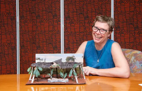 A Caucasian woman sits smiling at a table. She is wearing a blue sleeveless dress. She has one of her architectural design models in front of her.