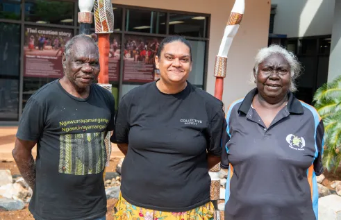 Three people stand looking at camera. And Aboriginal man is on the left wearing a black t-shirt. There is a woman in the middle and an elderly woman on the right. They all have warm, welcoming faces.