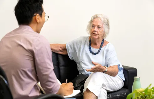 Person who is taking notes is facing an older person who is talking. They are both sitting down on a couch in a small consultation room.