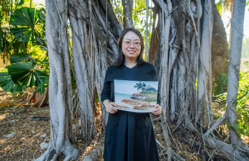 Young woman stands in front of native Australian trees on the Casuarina Campus in Darwin. She wears a black dress and cardigan and holds one of her watercolour pieces.