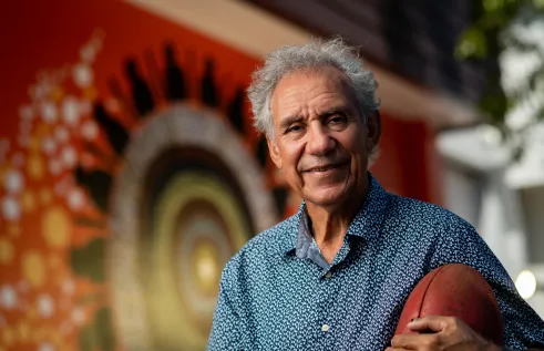 An Aboriginal man with greying hair and a warm smile holds a football while looking at the camera. He is standing in front of a First Nations artwork.