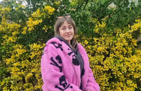 Young woman sits in front of wattle tree with yellow flowering. She wears a pink fluffy coat. 