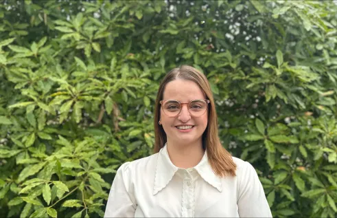 A young Caucasian woman stands in front of green foliage. She is looking directly at the camera and smiles. She is wearing a white blouse and glasses. 