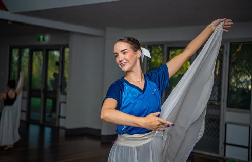 A young woman stands in a ballet pose. Her left arm is raised and holding a tulle skirt. 