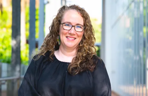 A woman with long curly brown hair and glasses is smiling outdoors. She is wearing a loose black blouse and colorful earrings, standing in a bright walkway with greenery and tall pillars in the background.
