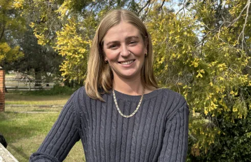 Young Caucasian woman standing outdoors looking at camera with a smile. She is wearing a grey jumper. 