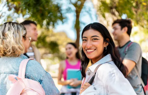 A young woman with brown hair smiles at the camera. She is wearing a white collared shirt. Next to her is a group of people engaged in discussion. They are standing outside. 
