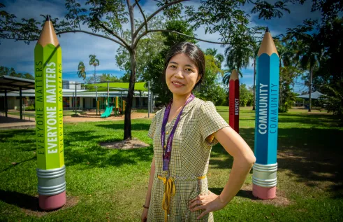 A young Chinese woman stands outside surrounded by larger than life pencils in vibrant colours. The pencils are as tall as she is and have words on them like 'everyone matters,' 'community,' and 'respect.'