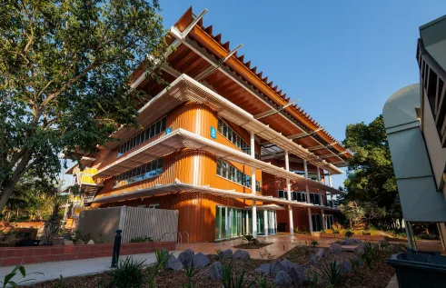 A contemporary three-storey university building with bright orange cladding, wide glass windows, and layered white verandas. The structure features striking angled roof panels for shade, with a blue number 8 sign marking the building. The foreground shows landscaped garden beds with rocks and native plants, and a clear blue sky forms the backdrop.