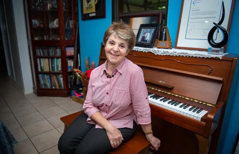 A smiling woman sits at a piano in her home, wearing a pink button-up shirt and dark pants. Behind her on the piano are framed photos, certificates, and music-related memorabilia, with a bookshelf visible in the background.