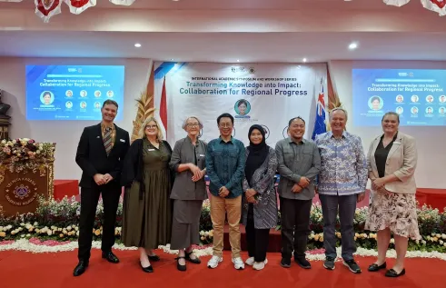 A group of eight people stand on a red-carpeted stage in front of a large conference backdrop that reads “Transforming Knowledge into Impact: Collaboration for Regional Progress.” They are smiling and posing for a photo after the event. The stage is decorated with flower arrangements, and banners of Indonesia and Australia are displayed. Two large screens project the event title and speaker details on either side of the stage.