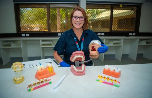 A woman wearing glasses and blue gloves smiles while seated at a dental training workstation. She holds a dental mannequin head with an open mouth, surrounded by large tooth and gum models, a giant toothbrush, and dental tools on the bench in front of her.