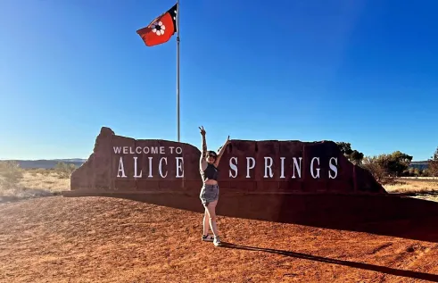 A person in shorts and a crop top stands in front of a large red rock sign that reads "Welcome to Alice Springs," raising both arms in a celebratory pose. Above the sign, a red flag with a white paw print design waves against a clear blue sky. The surrounding landscape is flat and dry with scattered trees and low hills in the background.