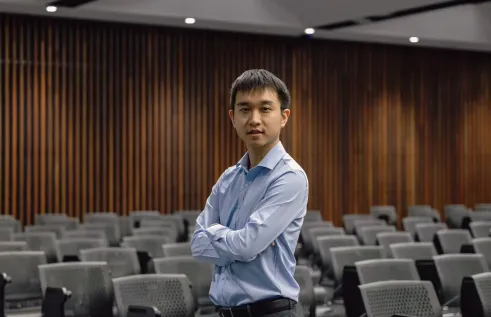 A man in a light blue dress shirt stands with arms folded in the middle of an empty lecture theatre. He faces the camera confidently, with rows of grey chairs behind him and vertical wooden wall panels in the background.