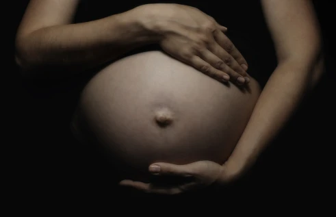 Close-up of a pregnant belly with two hands gently cradling it, one resting above and one below, against a dark background.