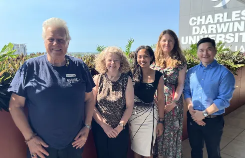 Five people stand together smiling on a sunny balcony at Charles Darwin University. The group includes three women and two men, dressed in smart casual attire, with lush green plants and a clear blue sky in the background. The CDU logo is visible on the building wall behind them.