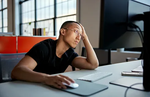 A person wearing glasses and a black t-shirt sits at a desk in front of a computer, resting their head on one hand while using a mouse with the other. They appear tired or frustrated, gazing off into the distance in a modern office with large windows and natural light.
