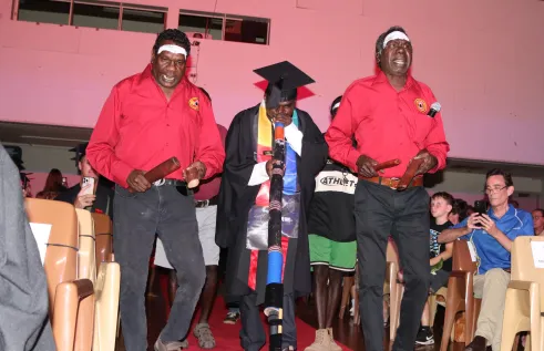 two TOs walk down read carpet with one graduate playing yidaki