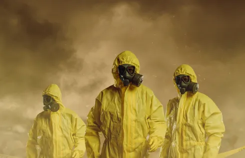 three people wearing full-body yellow hazmat suits and black gas masks. They are standing in a smoky, dusty, or foggy environment with a dark brownish sky, giving the scene an intense, possibly hazardous or post-apocalyptic feel. Yellow caution tape runs across the lower part of the image, suggesting a restricted or contaminated area.