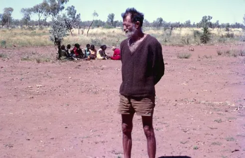 Man standing in a dirt area in the bus, with Indigenous people sitting behind him under a small tree