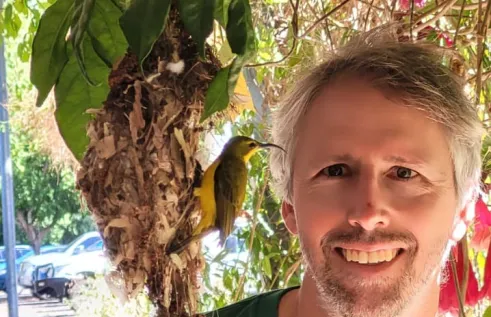 Golo Mauer head and shoulders, looking at small bird with long curved beak. yellow chest, and brown wings, perched on a nest hanging from leaves under a tree