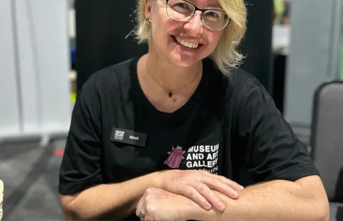 Dr Kirsti Abbott, wearing glasses, in black t-shirt with Museum and Art Gallery of the Northern TerritoryT logo on front, leaning on display case of insects