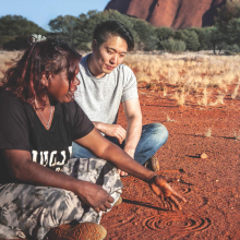 Indigenous woman explaining a figure drawn in the dirt to a man who is sitting with her and listening