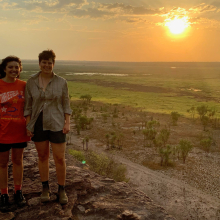 Two people standing on high rock with green plain in background and sun low in sky