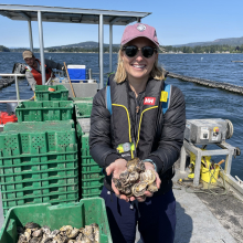 Samantha Nowland, wearing sunglasses and a cap, standing on a boat on water, holding a quantity of oysters in both hands. A person is in the background operating the boat.
