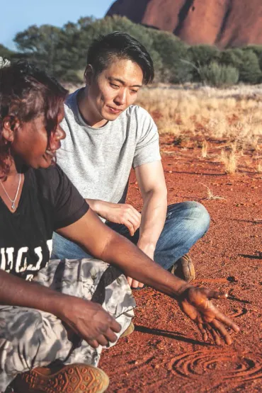 Indigenous woman explaining a figure drawn in the dirt to a man who is sitting with her and listening