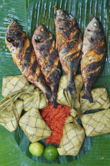 Cooked fish with limes, chilli paste and small parcels of rice on a bed coconut leaves