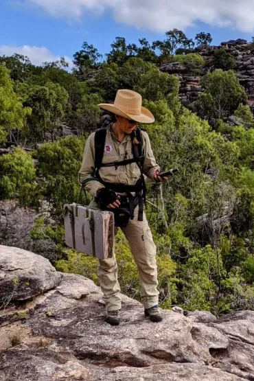 Person wearing hat standing on rocks looking at GPS, with trees in background