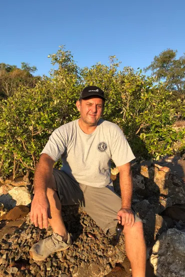 Dr Benjamin Brown sitting on rock in sun with leafy bush behind him