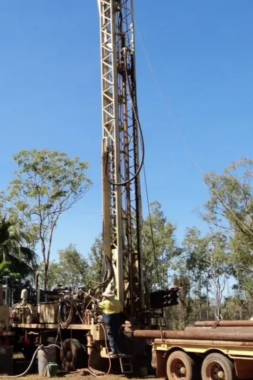 Tall drill rig on back of truck, with person in hard had and hi-vis working at the bottom, with trees in the background
