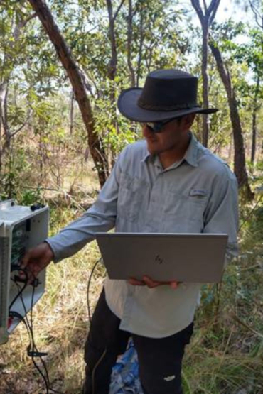 Dr Francesco Ulloa Cedamanos standing in grassy savannah, holding a laptop and adjusting some equipment in a box