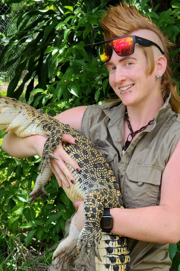 Person with mohawk hairstyle, with sunglasses on their forehead, holding crocodile about the size of a medium sized dog, with green leafy background