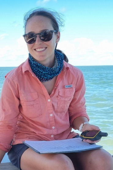 Natalie Robson wearing an orange shirt and sunglasses, holding a GPS and a clipboard, apparently sitting on a boat, with sea in the background