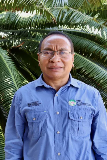 Acacio da Costa Guterres from waist up, wearing blue shirt with CDU and RIEL logos, with leaves in the background