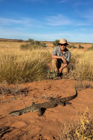 Dr Nicholas Wu, wearing a hat and sunglasses, crouching near some clumps of grass, with a large lizaard on bare soil in the foreground