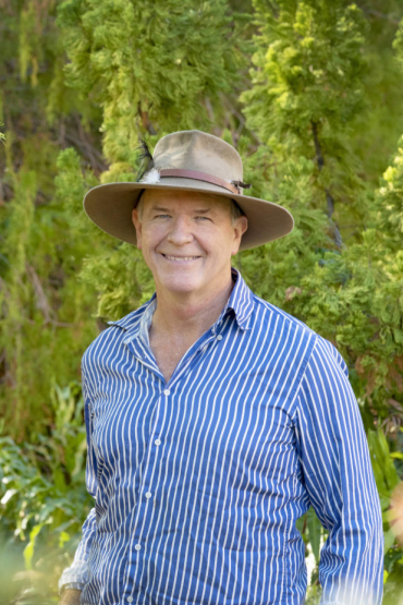 Professor Stephen Garnett head and shoulders, wearing blue and white striped shirt and an Akubra hat, with green leafy vegetation in background