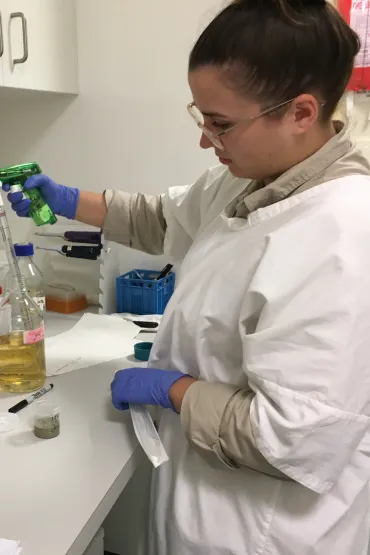 A person wearing glasses and a white lab coat standing at a bench, holding a pipette in a clear glass bottle containing yellow liquid