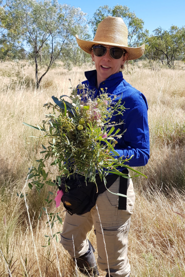 Doctor Donna Lewis wearing hat and sunglasses, holding a bunch of native flowers and leaves, standing in long dry grass with trees in background