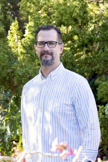 Dr Keller Kopf with a beard and glasses, wearing a shirt with vertical light blue stripes, with dense green foliage behind
