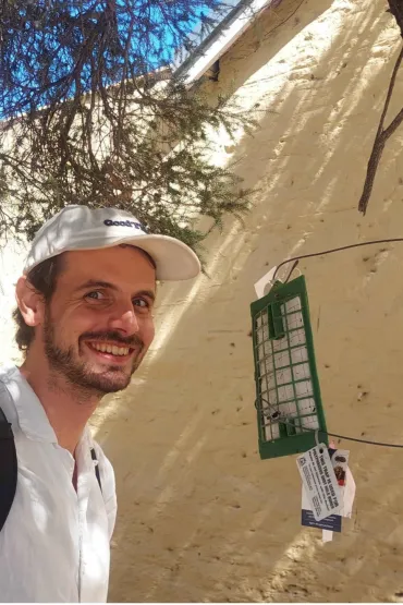 Andrew Coates, head and chest, on the left of the photo, in a white shirt and a white cap, smiling at the camera, in front of a white wall, with tree branches above his head. A bug trap attached by wires to a tree trunk which is just visible at the right of the image