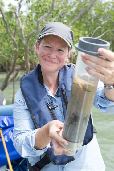 Julia Fortune holding a clear container with brown water and mud inside, with water and mangroves in the background