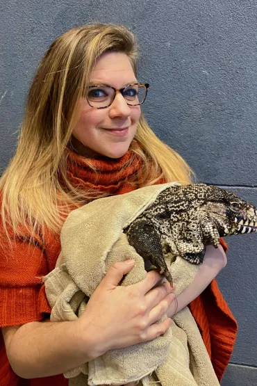 Sarah Snyder dressed in a red/brown jumper, holding a large black and white lizard partly wrapped in a towel, with its tongue sticking out