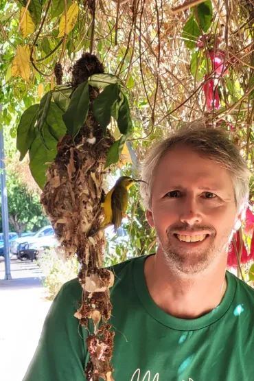 Golo Mauer under a tree, wearing a green t-shirt, looking at small bird with long curved beak. yellow chest, and brown wings, perched on a messy looking nest hanging from leaves