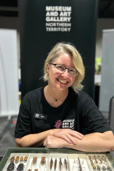 Dr Kirsti Abbott, wearing glasses, in black t-shirt with Museum and Art Gallery of the Northern TerritoryT logo on front, leaning on display case of insects. In the background is a black banner with "Museum and Art Gallery of the Northern Territory" written on it